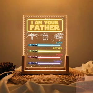 May include: A lighted acrylic sign with the text "I AM YOUR FATHER" and illustrations of spaceships and a helmet. Below are four lightsaber designs with names: Luke, Petter, John, and Tommy. The sign is set in a wooden frame.