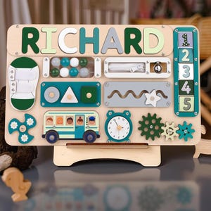 May include: Wooden activity board with the name "RICHARD" in green, white, and gray letters. Features include a zipper, abacus, clock, gears, and a school bus with children. The board is designed for educational play.