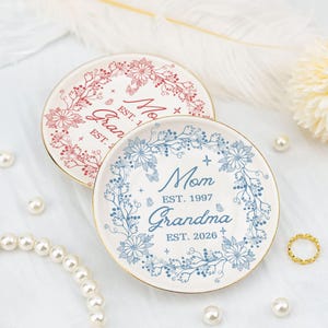 Könnte beinhalten: Zwei dekorative Keramikplatten mit floralen Designs und goldenen Rändern. Eine Platte zeigt einen roten Blumenkranz mit den Worten "Mom" und "Grandma". Die andere Platte hat einen blauen Blumenkranz mit den Worten "Mom" und "Grandma" und den Jahren 1997 und 2026.