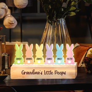 Könnte beinhalten: Ein Holzschild mit dem Text "Grandma's Little Peeps" und sechs leuchtenden Hasenfiguren mit den Namen "Emily", "Emma", "James", "Linda", "Max" und "Betty".