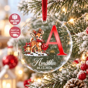 Könnte beinhalten: Ein runder, klarer, facettierter Glas-Weihnachtsbaumschmuck, der an einem roten Band hängt. Er zeigt ein Cartoon-Rehkitz mit einem rot karierten Schal, einen großen roten Buchstaben A, den Namen Amatha und das Datum 10.12.2026.