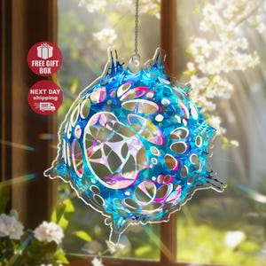 May include: A hanging, abstract suncatcher with a complex, geometric design. The suncatcher is primarily blue with iridescent accents, and features a series of cut-out shapes. It hangs from a silver chain, and is displayed in front of a window.