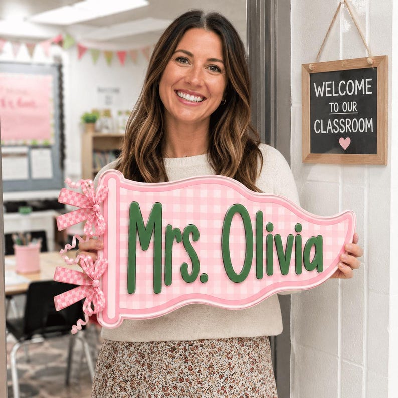 May include: A pennant-shaped sign featuring a light pink and white gingham pattern with the text Mrs. Olivia in raised dark green lettering. The left side of the sign is decorated with two pink and white checkered bows with curly ribbon accents.