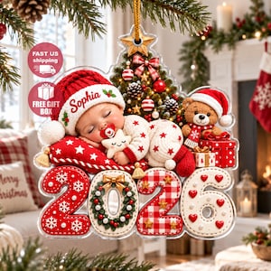 Könnte beinhalten: Ein Weihnachtsbaumschmuck mit einem schlafenden Baby in einer roten Mütze mit dem Namen Sophia, einem Teddybären und der Jahreszahl 2026. Das Design zeigt ein rot-weißes Farbschema mit Sternen, einem Kranz und Geschenkboxen an einer goldenen Schnur.
