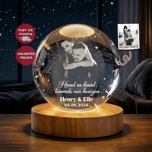 Könnte beinhalten: Ein kugelförmiges Glasornament auf einem runden Holzsockel mit einem lasergravierten Foto eines Paares im Inneren. Das Glas enthält den eingravierten Text "Hand in hand, towards our horizon," "Henry & Elle," und das Datum "06.06.2026".