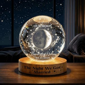May include: A clear glass sphere lamp featuring an etched moon and star constellation design. The text reads OUR MOON, Brian & Hellan, 08.15.2020. The wooden base is engraved with the text The Night We Got Married and decorated with small stars.