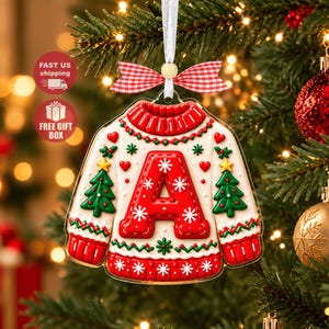 Könnte beinhalten: Ein flacher, pulloverförmiger Weihnachtsbaumanhänger mit weißer Basis, rotem Saum, grünen Tannenbäumen und einem großen roten Buchstaben A in der Mitte. Der Anhänger ist mit einem rot-weiß karierten Band verziert und hängt an einem weißen Satinband.