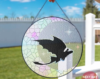 Puede incluir: Obra de arte circular de vidriera con una silueta de dragón negro sobre un fondo geométrico colorido. La pieza está suspendida por una cadena. El fondo muestra un cielo azul y hierba verde.