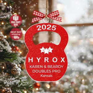 May include: A red glittered kettlebell-shaped Christmas ornament with the year 2025 in white. The ornament has the text "HYROX KAREN & BEARDY DOUBLES PRO Kansas" and is adorned with a red and white bow.
