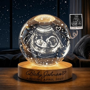 May include: A spherical glass ornament featuring an etched ultrasound image of a fetus, surrounded by engraved stars and hearts. The glass sphere rests on a circular wooden base with the text Baby Johnson ARIVING APRIL 2027 engraved on it.