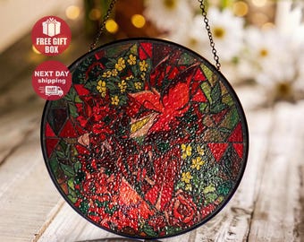 May include: Round stained glass window art with a floral design in red, green, and yellow. The artwork is framed in black and hangs from a chain. The design features roses and other flowers.