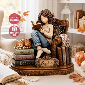 Könnte beinhalten: Eine Figur einer Person mit langem braunem Haar, die in einem braunen Sessel sitzt und ein Buch liest. Eine orangefarbene getigerte Katze schläft auf drei Büchern mit den Aufschriften Just A Girl, Who Loves und Books. Ein Schild trägt den Namen Sophia.