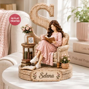 Könnte beinhalten: Eine dekorative Figur, die eine Person mit langem braunem Haar zeigt, die in einem Sessel sitzt und ein Buch liest, bekleidet mit einem rosa Kleid und weißen Turnschuhen. Der Sockel trägt den Namen Selena und hinter dem Sessel steht ein großer Buchstabe S. Auf einem kleinen Tisch stehen eine Vase mit rosa Blumen, ein Schild mit der Aufschrift Just one more chapter und gestapelte Bücher.