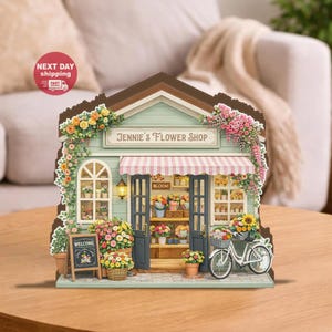 Könnte beinhalten: Ein Miniaturmodell eines Blumengeschäfts mit hellblauer Fassade, einem rosa-weiß gestreiften Vordach und dem Text "Jennie's Flower Shop". Das Geschäft ist mit bunten Blumen, einem Fahrrad und einem "Welcome"-Schild geschmückt.