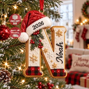 Könnte beinhalten: Ein Weihnachtsanhänger in Form des Buchstabens N mit einer roten Glitzer-Weihnachtsmannmütze mit der Jahreszahl 2026, grünen Stechpalmenblättern mit roten Beeren und goldenen Schneeflocken. Die Basis ist weiß mit rot-schwarzem Karomuster und dem Namen Noah.