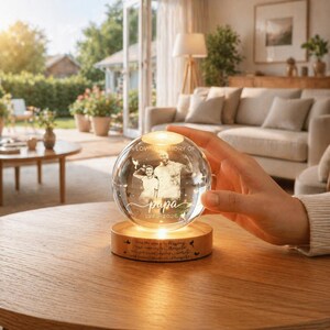 May include: A clear glass sphere with a photo of a man and a child, resting on a wooden base. The sphere is illuminated from within, and the base has engraved text. The text reads "In loving memory of Papa 1940-2026".