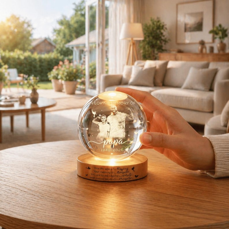 May include: A clear glass sphere with a photo of a man and a child, resting on a wooden base. The sphere is illuminated from within, and the base has engraved text. The text reads "In loving memory of Papa 1940-2026".
