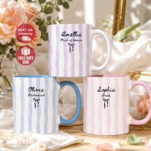 Könnte beinhalten: Drei Keramiktassen mit vertikalen, wellenförmigen Streifen in Lavendel, Hellblau und Rosa. Jede Tasse zeigt einen Namen, eine Rolle und eine kleine schwarze Schleife. Die lavendelfarbene Tasse trägt Amellia Maid of Honor, die blaue Olivia Bridesmaid und die rosa Sophia Bride.