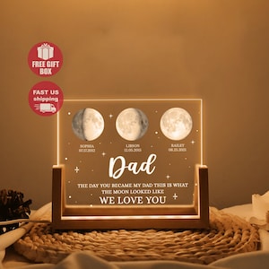 Könnte beinhalten: Ein personalisiertes LED-Nachtlicht mit Holzsockel. Das Licht zeigt drei Mondphasenbilder und das Wort "Dad" in der Mitte. Der Text enthält Namen und Daten mit dem Satz "An dem Tag, an dem du mein Papa wurdest, so sah der Mond aus. Wir lieben dich."