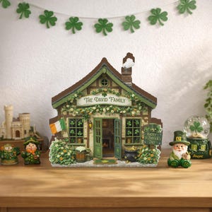 May include: A detailed miniature house with green trim and a brown roof, adorned with floral accents and an Irish flag. The sign reads "The David Family" and "Happy St. Patrick's Day". Decorative figurines and a shamrock garland complete the festive scene.