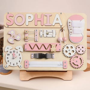 May include: A pink and wooden activity board with the name "Sophia" spelled out in pink letters. The board has a variety of activities for children to explore, including a clock, a zipper, a buckle, a maze, gears, and a shoe with laces.