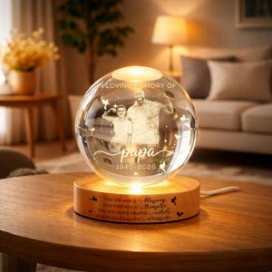 May include: A clear glass sphere with a photo of two people inside, resting on a wooden base. The sphere is illuminated from below, with the words "IN LOVING MEMORY OF" and "papa 1940-2026" etched on the surface. The wooden base has an inscription.