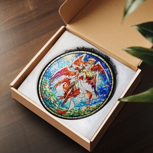 May include: A round stained glass art piece featuring a colorful illustration of a character with red wings and a hat, set against a blue and floral background. The artwork is displayed in a cardboard box with white padding.