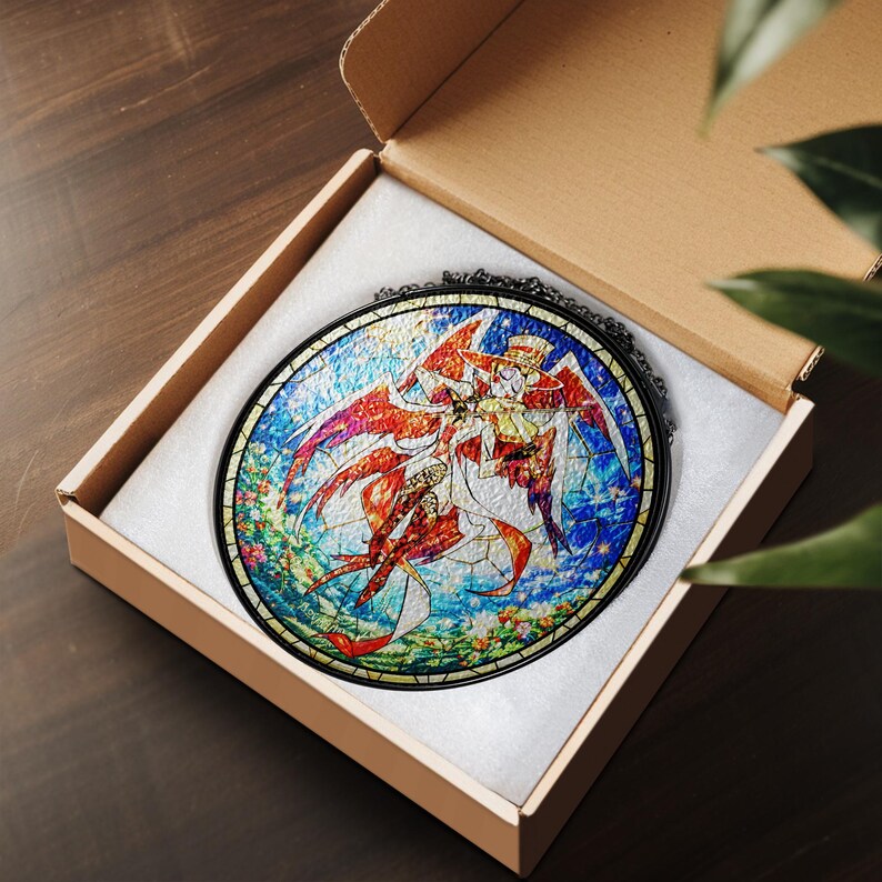 May include: A round stained glass art piece featuring a colorful illustration of a character with red wings and a hat, set against a blue and floral background. The artwork is displayed in a cardboard box with white padding.
