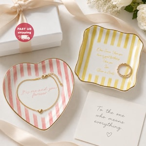 Könnte beinhalten: Eine herzförmige Keramikschale mit rosa Streifen und dem Text 'It's me and you forever' mit einem goldenen Herzarmband, daneben eine quadratische gelb gestreifte Schale mit goldenem Text und Ring sowie eine Karte mit 'To the one who means everything'.