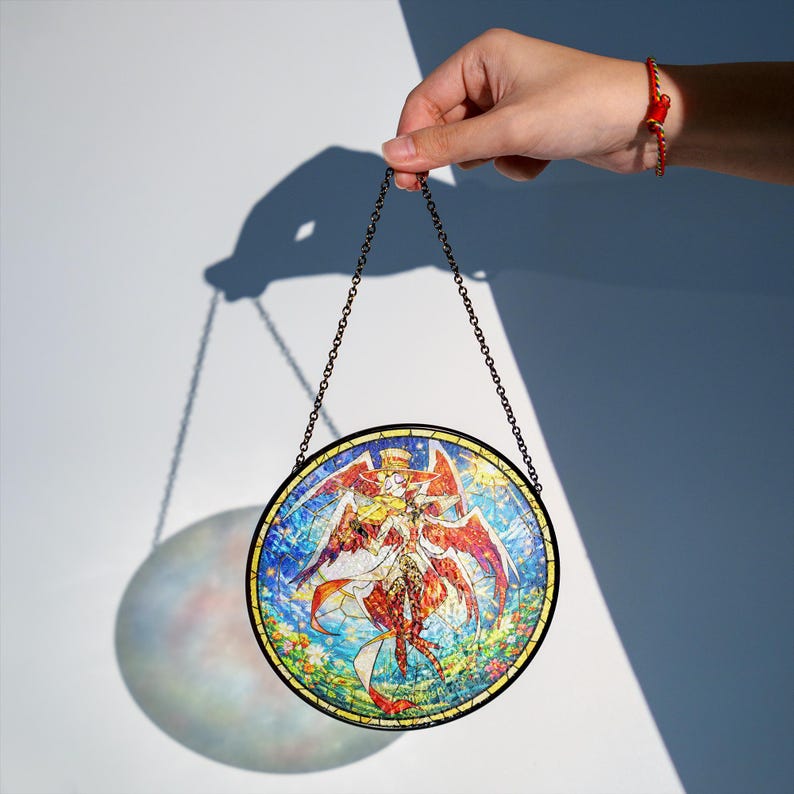 May include: A round stained glass panel featuring a detailed angel design with red and gold wings, held by a chain. The artwork has a black border and is set against a white background. The image is illuminated, casting a shadow.