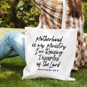 May include: White canvas tote bag with black text that reads "Motherhood is my Ministry I'm Raising Disciples of the Lord Proverbs 22:6".