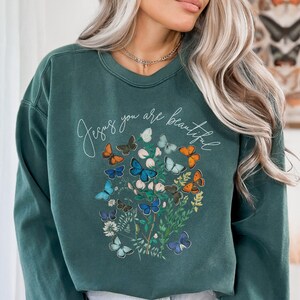Könnte beinhalten: Ein dunkelgrüner Pullover mit einem Schmetterlingsmotiv und dem Text "Jesus you are beautiful".