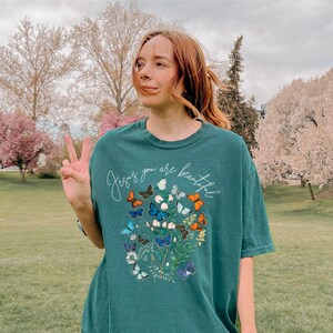 Könnte beinhalten: Ein blaugrünes T-Shirt mit einem Aufdruck von Schmetterlingen und Blumen. Der Text "Jesus you are beautiful" ist auf das Shirt gedruckt.