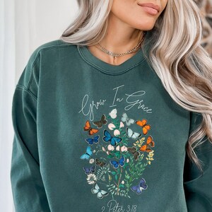 Könnte beinhalten: Ein dunkelgrüner Pullover mit einem weißen Aufdruck von Schmetterlingen und Blumen. Der Text "Grow in Grace" und "2 Peter 3:18" ist auf dem Pullover aufgedruckt.