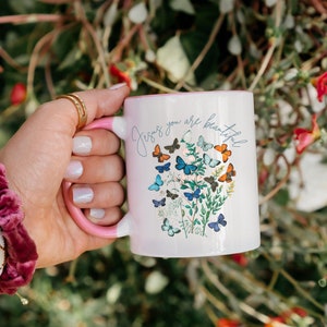 Może przedstawiać: Biały kubek ceramiczny z różowym rantem i uchwytem. Kubek zdobi kwiatowy wzór z motylami i tekstem "Jesus you are beautiful".