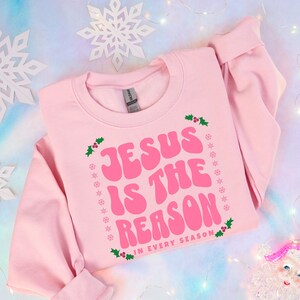 Könnte beinhalten: Rosafarbener Pullover mit dem Text "Jesus ist der Grund in jeder Jahreszeit" in Rosa mit grünen Stechpalmen-Akzenten.
