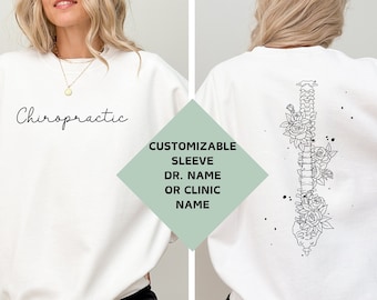 Op de afbeelding: Witte trui met het woord "Chiropractic" op de voorkant en een bloemig ruggengraatontwerp op de achterkant. De mouw heeft ruimte voor een aanpasbare naam of klinieknaam.