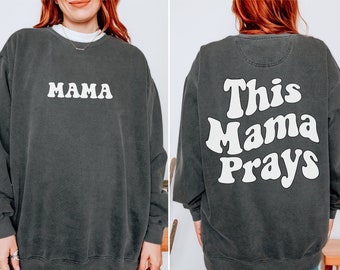 Könnte beinhalten: Ein schwarzer Pullover mit dem Text "MAMA" auf der Vorderseite und "This Mama Prays" auf der Rückseite in einer Retro-Schrift.