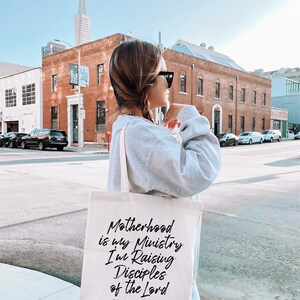 May include: A white canvas tote bag with black text that reads "Motherhood is my Ministry I'm Raising Disciples of the Lord Proverbs 22:6".