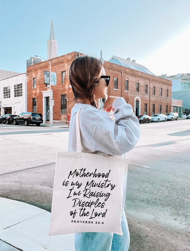 May include: A white canvas tote bag with black text that reads "Motherhood is my Ministry I'm Raising Disciples of the Lord Proverbs 22:6".