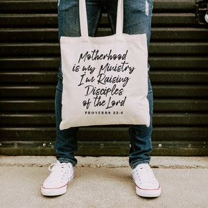 May include: A white canvas tote bag with the text "Motherhood is my Ministry I'm Raising Disciples of the Lord Proverbs 22:6" printed in black.