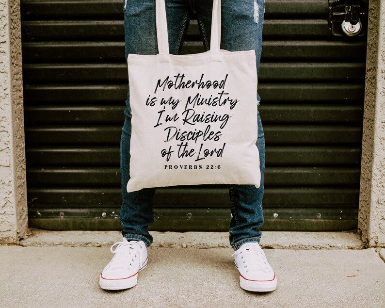 May include: A white canvas tote bag with the text "Motherhood is my Ministry I'm Raising Disciples of the Lord Proverbs 22:6" printed in black.
