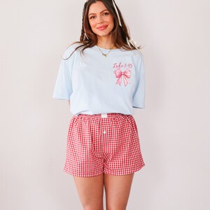 May include: A light blue t-shirt with a pink bow and the text "Luke 1:45" printed on the front. The person is wearing red and white gingham shorts and black cowboy boots.