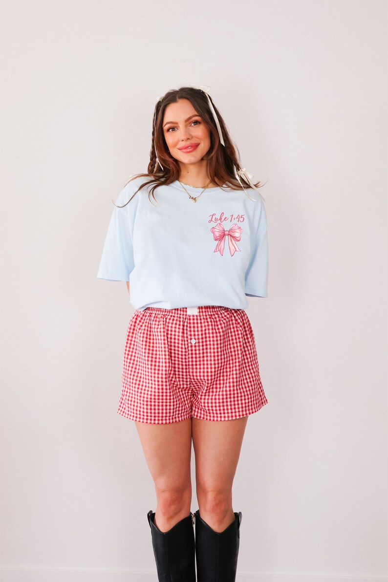 May include: A light blue t-shirt with a pink bow and the text "Luke 1:45" printed on the front. The person is wearing red and white gingham shorts and black cowboy boots.