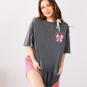 May include: An oversized charcoal grey t-shirt with a pink bow and the text "Luke 1:45" printed on the front. The model is wearing the t-shirt with red and white gingham shorts and black boots.
