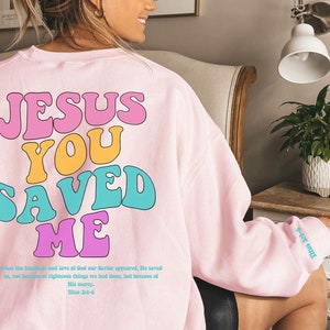 Könnte beinhalten: Rosafarbener Pullover mit dem Text "JESUS YOU SAVED ME" in bunten Retro-Lettern. Der Text "Titus 3:4-5" ist unter dem Haupttext gedruckt.