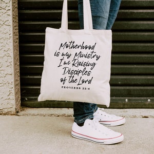 May include: A white canvas tote bag with black text that reads "Motherhood is my Ministry I'm Raising Disciples of the Lord Proverbs 22:6".