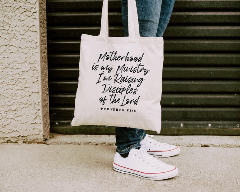 May include: A white canvas tote bag with black text that reads "Motherhood is my Ministry I'm Raising Disciples of the Lord Proverbs 22:6".