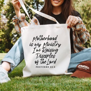 May include: A white tote bag with black text that reads "Motherhood is my Ministry I'm Raising Disciples of the Lord Proverbs 22:6".