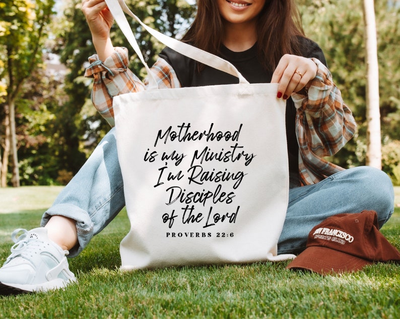 May include: A white tote bag with black text that reads "Motherhood is my Ministry I'm Raising Disciples of the Lord Proverbs 22:6".